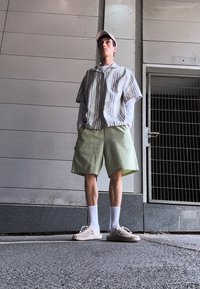 Camisa de manga corta a rayas en beige y blanco, pantalones cortos verdes texturizados, calcetines blancos, sandalias beige de dedo abierto, de pie contra una pared gris.
