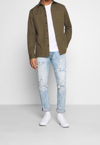 Camisa de botones color verde oliva, camiseta blanca debajo, jeans desgastados azul claro y zapatillas blancas. La camisa tiene dos bolsillos en el pecho.