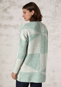 Lichtgrijze cardigan met teal geometrisch patroon, lange mouwen en een ronde halslijn. Zachte, gestructureerde breimateriaal. Zijsplitten aan de onderkant.