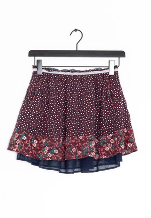 Mørkerød lagdelt nederdel med små hvide prikker og blomsterpatterkant hængende på en sort bøjle mod en hvid baggrund.