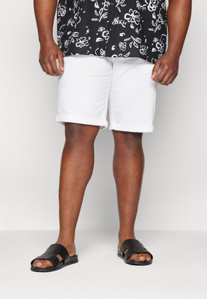 Weiße Baumwollshorts mit umgeschlagenem Saum, kombiniert mit schwarzen strukturierten Sandalen. Der Hintergrund ist einfarbig grau. Fokus auf dem Unterkörper.