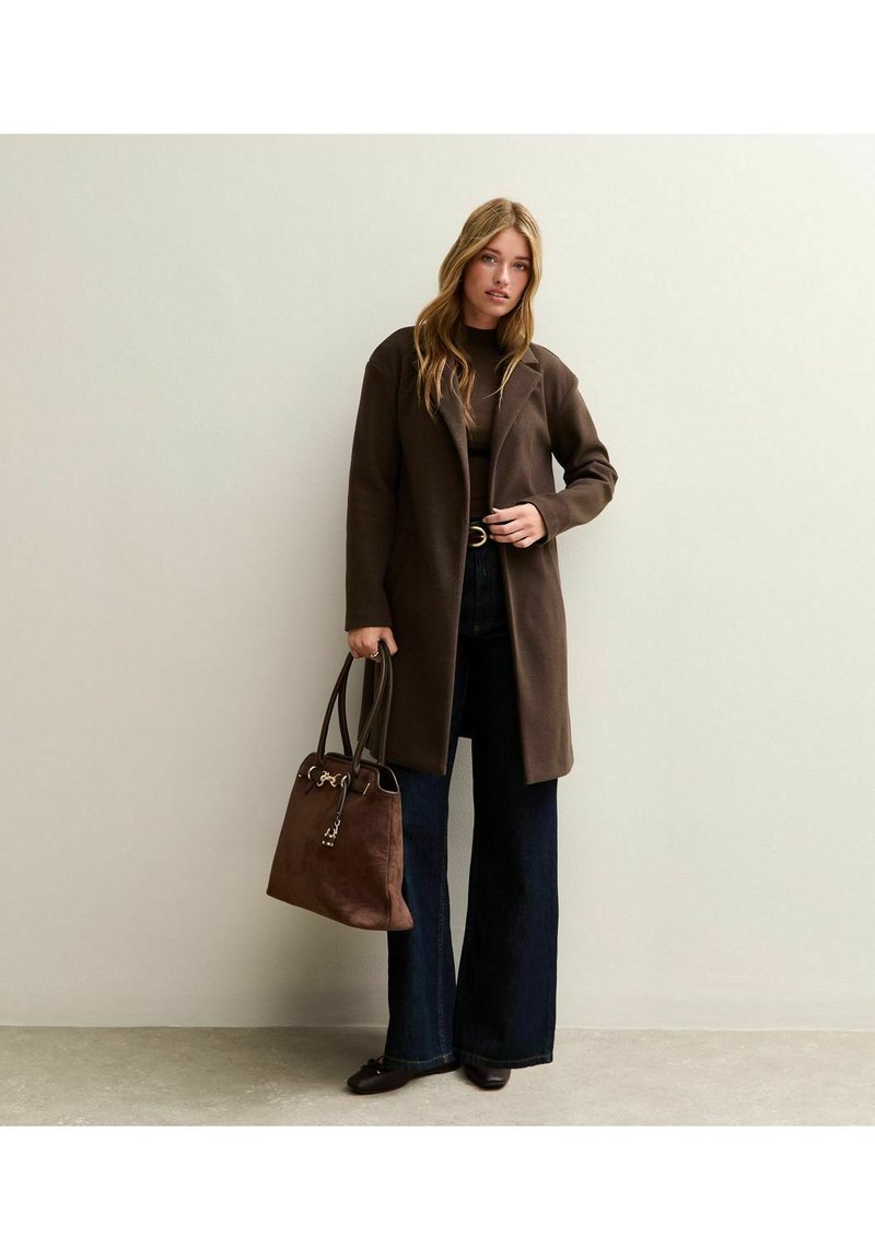 Abrigo de mezcla de lana marrón con un cuello redondeado, llevado sobre una blusa oscura y combinado con jeans acampanados y un bolso marrón. Textura suave.