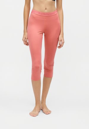 Mujer con leggings de coral rosa de tres cuartos con cintura "Peak Performance", de pie descalza sobre un fondo claro.