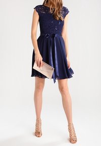 Robe de soirée marine avec un corsage en dentelle, des manches courtes, et une ceinture en satin, accompagnée d'une pochette. Portée avec des talons nus à lanières, dotée d'une jupe évasée.