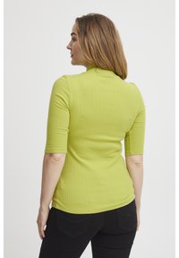 Kortärmad, ribbad topp i ljus limegrön, med hög halsringning och figurnära design. Svarta byxor delvis synliga nedtill.