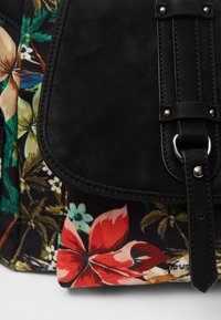 Czarna skórzana klapka z klamrą na plecaku z tkaniny w kwiaty, zdobionym wzorami tropikalnych kwiatów i liści w kolorach czerwonym, beżowym i zielonym.
