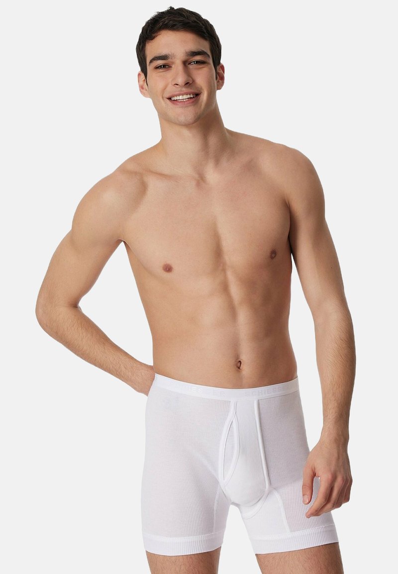 White cotton boxers with a ribbed texture, featuring a front opening and a soft waistband. Model poses with a neutral expression.