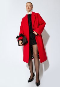 Abrigo rojo de gran tamaño con solapas anchas, combinado con un vestido negro texturizado y una falda negra con lunares, sosteniendo un bolso negro.