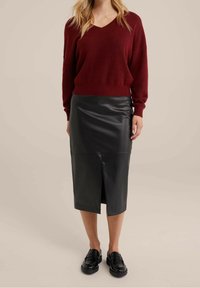 Frau trägt einen bordeauxroten V-Ausschnitt-Pullover, schwarzen Leder-Midirock mit Frontschlitz und schwarze Loafers, steht vor einem schlichten beigen Hintergrund.