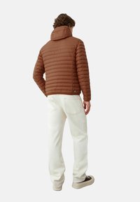 Bruine pufferjas met horizontale stiksels en een capuchon, gecombineerd met witte jeans en donkere sneakers, gezien vanaf de achterkant.
