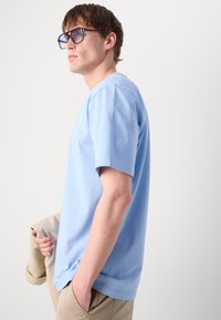 Ανοιχτού μπλε βαμβακερό T-shirt κοντό μανίκι με στρογγυλή λαιμόκοψη, απαλό υφή και χαλαρή εφαρμογή. Το μοντέλο κρατά ένα μπεζ αντικείμενο πάνω από τον ώμο του.