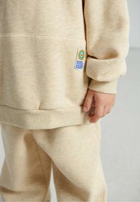 Ensemble sweat-shirt et pantalon beige en tissu doux. Comprend une étiquette cousue avec un motif circulaire, des poignets côtelés et une coupe décontractée.