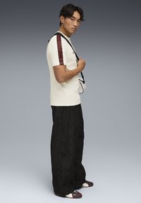 Tricou crem cu accente roșii pe mâneci, pantaloni negri supradimensionați și pantofi multicolori; modelul stă de profil împotriva unui fundal gradient.