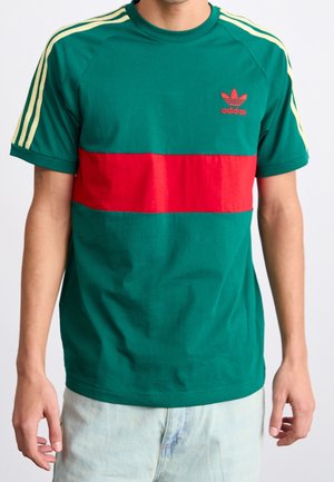 T-shirt en coton vert avec une bande horizontale rouge, trois bandes jaunes sur les épaules et un petit logo Adidas rouge sur la poitrine.