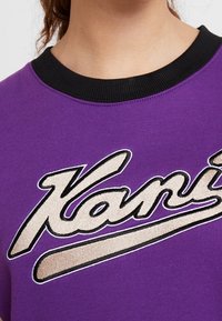 Camiseta de algodón morada con cuello acanalado negro, que presenta un gran logo bordado "Kani" en color crema y negro.