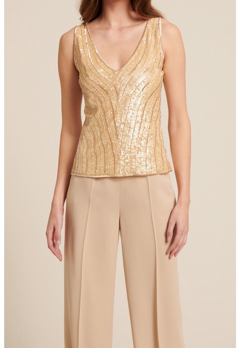 Kvinde iført en ærmeløs top med guldpalietter og v-hals samt brede beige bukser foran en ensfarvet baggrund.