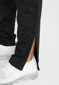 Svarta sportbyxor med sidoförslutning med dragkedja och meshdetaljer vid anklarna, kombinerade med vita sportskor.