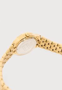 Χρυσής απόχρωσης μεταλλικό μπρασελέ ρολογιού με κλειστό κούμπωμα και το λογότυπο Tommy Hilfiger ορατό στην πίσω πλευρά της κάσας του ρολογιού.