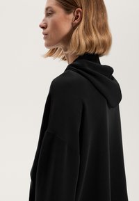 Felpa nera con cappuccio, dalla vestibilità ampia, caratterizzata da una texture morbida, spalle scese e dettagli di cucitura minimali. Vista laterale.