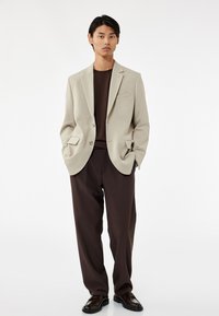 Lichtgrijze blazer met twee knopen, gecombineerd met een donkerbruine shirt en losse broeken. Gladde textuur en op maat gemaakte ontwerp, geaccentueerd met zakken.