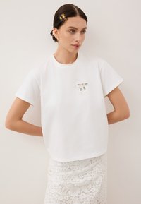 Witte katoenen T-shirt met een ronde hals, korte mouwen en twee zwart-witte labels op de voorkant. Gelaagd kantrokje zichtbaar eronder.