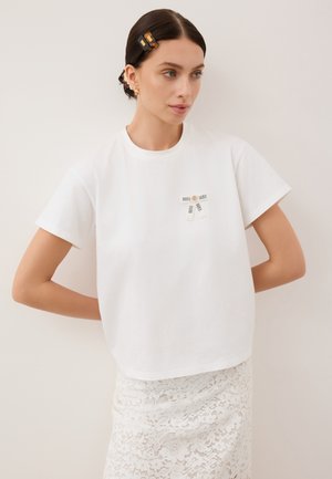 Witte katoenen T-shirt met een ronde hals, korte mouwen en twee zwart-witte labels op de voorkant. Gelaagd kantrokje zichtbaar eronder.