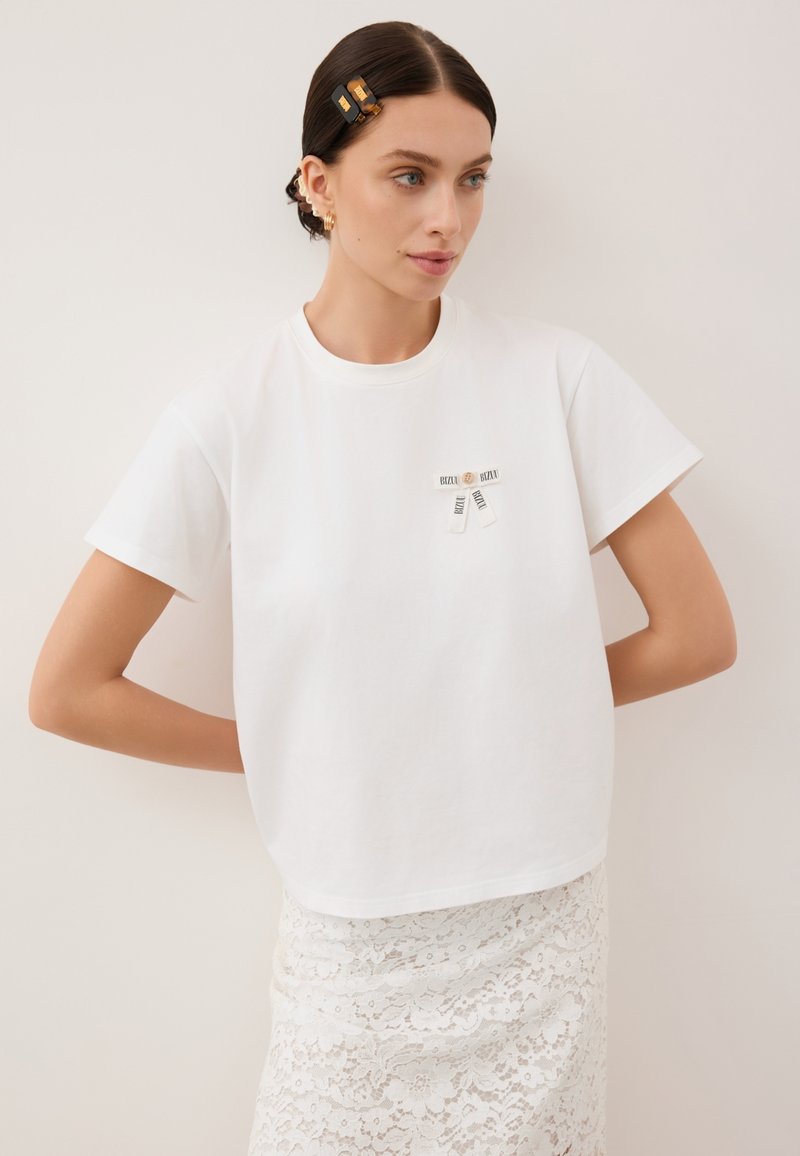 Witte katoenen T-shirt met een ronde hals, korte mouwen en twee zwart-witte labels op de voorkant. Gelaagd kantrokje zichtbaar eronder.