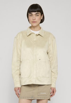 Mujer con cabello corto y oscuro que lleva una chaqueta beige abotonada y una falda beige a juego con bolsillos delanteros visibles y marca.