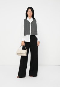 Kvinne iført hvit skjorte, grå stripete vest, svarte vide bukser, svarte sandaler, holder en beige håndveske.