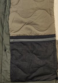 Die Innenseite einer gesteppte Jacke zeigt eine schwarze Netz-Reißverschlusstasche, olivgrüne Stoffkante und beiges Fleece-Futter auf der rechten Seite.