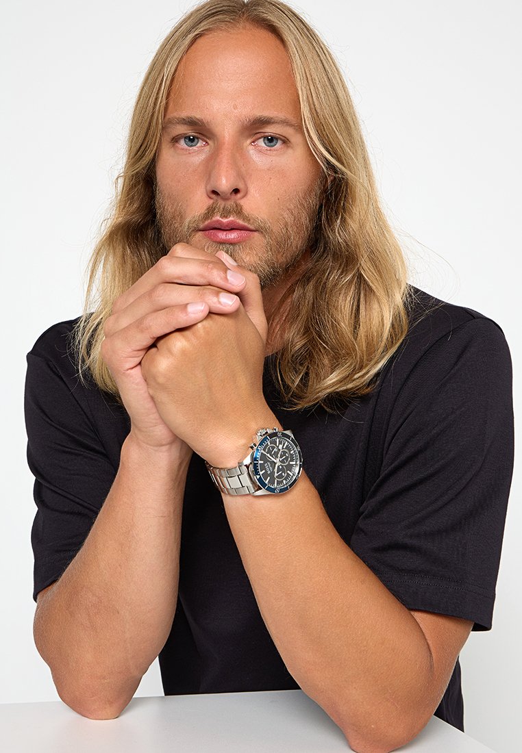 Reloj de acero inoxidable plateado con una esfera azul, características de cronógrafo y una correa metálica, llevado en una mano que descansa sobre unos brazos cruzados.