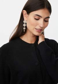 Černý pletený cardigan s kulatým výstřihem a velkými knoflíky. Modelka nosí třpytivé náušnice se květinovým motivem, které zdobí různé krystalové tvary.