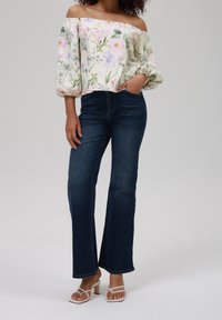 Blusa floreale off-the-shoulder con maniche a sbuffo, abbinata a jeans a campana blu scuro e sandali bianchi con tacco a blocco.