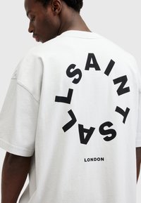 Hvit bomull T-skjorte med en bold svart sirkulær tekstdesign som sier "SAINT LONDON" på baksiden, med en avslappet passform og rund hals.