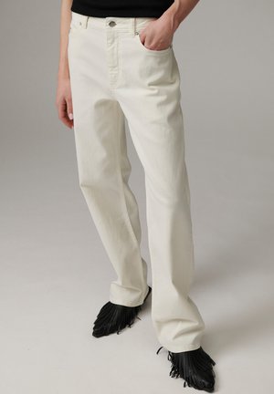 Personne portant un pantalon droit écru, une main dans la poche, associé à des chaussures noires à franges et un haut noir, debout sur un fond gris uni.