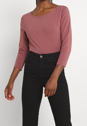 Osoba ubrana w bluzkę w kolorze mauve z rękawami do łokcia, włożoną do wysokich czarnych jeansów, stojąca na tle gładkiej białej ściany.