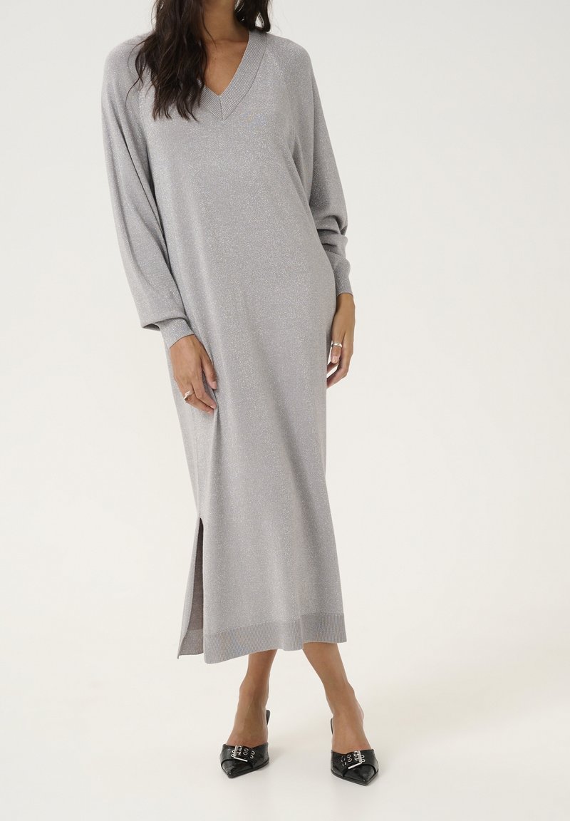 Femme portant une longue robe en maille gris clair à col en V, manches longues et fentes latérales, associée à des talons noirs à bout pointu.