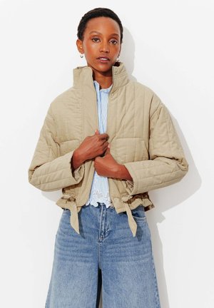 Femme ajustant une veste matelassée beige sur un chemisier à rayures bleues et un jean bleu ample sur fond blanc.