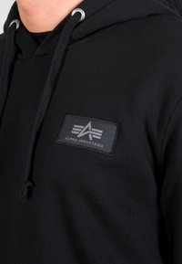 Dettaglio ravvicinato di una felpa nera con cappuccio, con una patch del logo Alpha Industries sul petto e lacci neri con occhielli in metallo.