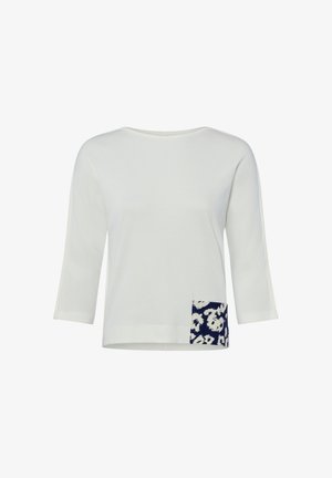 Blouse blanche à manches longues avec un col rond et un petit patch à imprimé animal bleu marine et blanc près de l'ourlet inférieur.