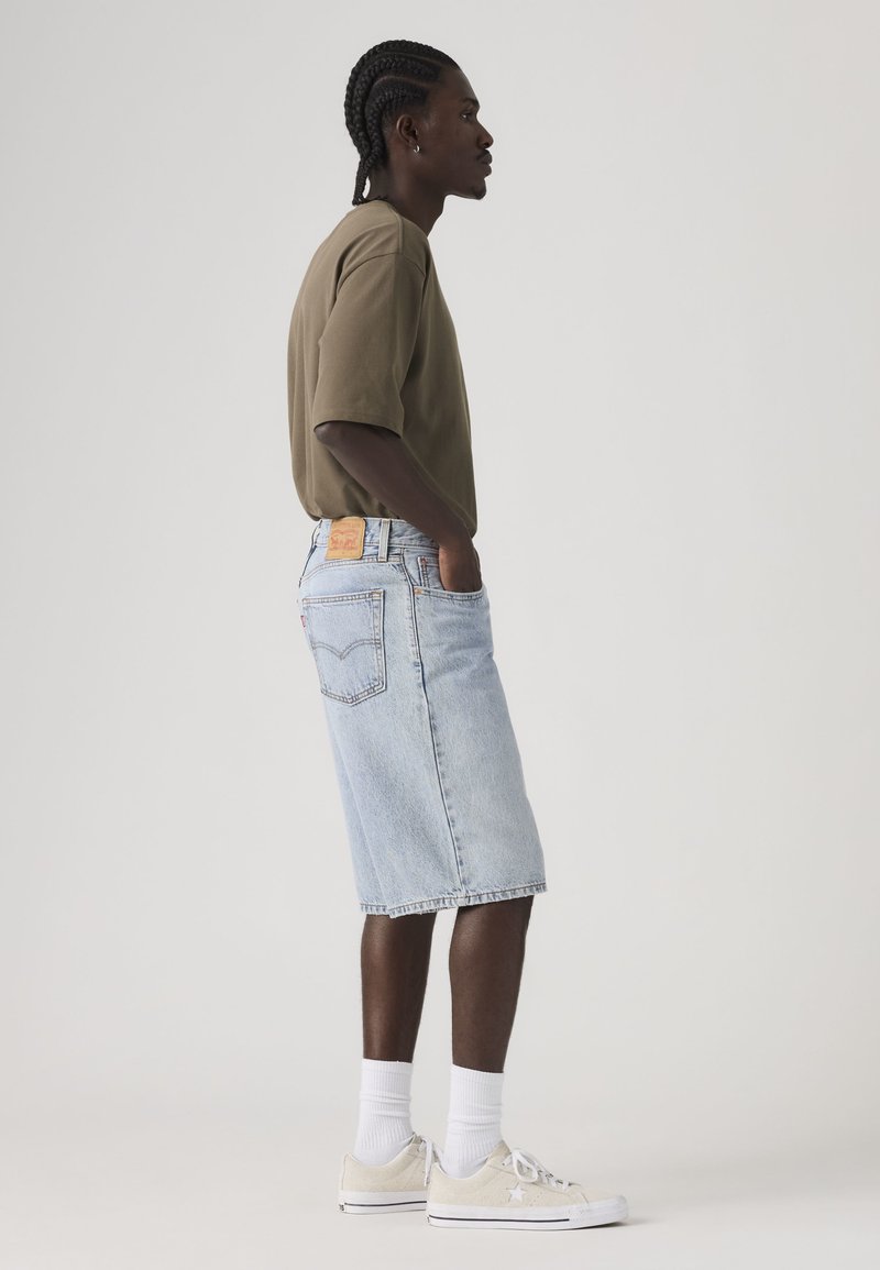 Junger Mann mit geflochtenem Haar, der ein braunes T-Shirt, hellblaue Jeans-Knielange Shorts, weiße Socken und cremefarbene Sneaker trägt, im Profil stehend.