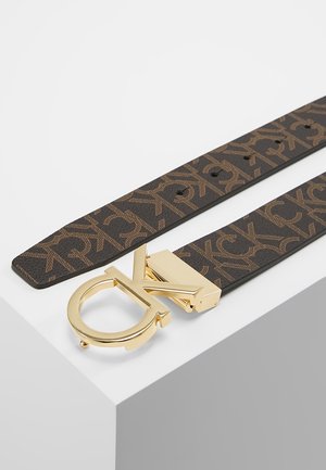 Brauner, gemusterter Gürtel mit goldfarbener, metallischer CK-Monogramm-Schnalle, flach auf einer weißen Oberfläche liegend.