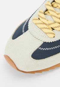 Suede beige sneakers med navy stoffdetaljer, gule stripete snørebånd og gummisåle. Har dekorativ søm og glatt tekstur.