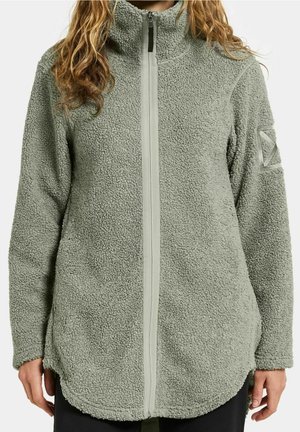 Persoon draagt een lichtgroene sherpa-fleece jas met rits en hoge kraag, met logo op de linker mouw, handen ontspannen langs het lichaam.