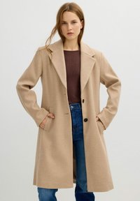 Abrigo de mezcla de lana color beige con grandes solapas, cierre de un solo botón, bolsillos frontales y mangas largas, llevado sobre una parte superior de canalé marrón y jeans azules.
