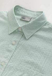 Chemise boutonnière texturée vert clair avec col pointu, trois boutons blancs et une poche poitrine, étiquetée "NEXT EDIT."