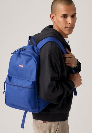 Blå ryggsekk laget av stoff, med en frontlomme, justerbare stropper og Levi's-logo. Plassert mot en nøytral bakgrunn.