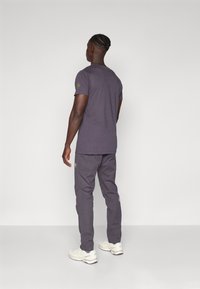 Γκρίζο βαμβακερό μπλουζάκι με κοντά μανίκια, σε συνδυασμό με αντίστοιχους γκρι παντελονιού. Σημαντική λεπτομέρεια λογότυπου στο μανίκι. Φορεμένο με λευκά αθλητικά παπούτσια.