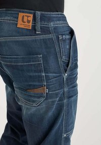 Jeans in denim blu scuro con un patch in pelle, numerose tasche, cuciture a contrasto e una finitura leggermente usurata.