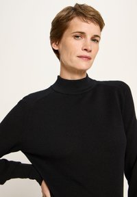 Frau mit kurzen braunen Haaren, die ein schwarzes, strukturiertes Langarmoberteil mit einem Stehkragen trägt und mit einer Hand auf der Hüfte vor einem schlichten Hintergrund posiert.